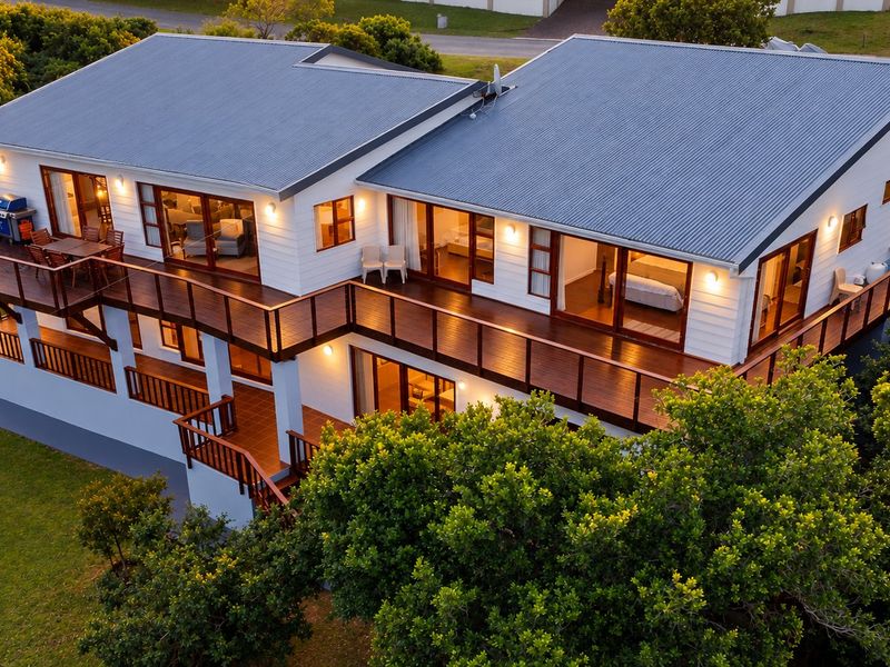 Aerial view of a modern house featuring a large wrap-around timber deck, corrugated metal roof, and extensive sliding glass doors overlooking a garden.