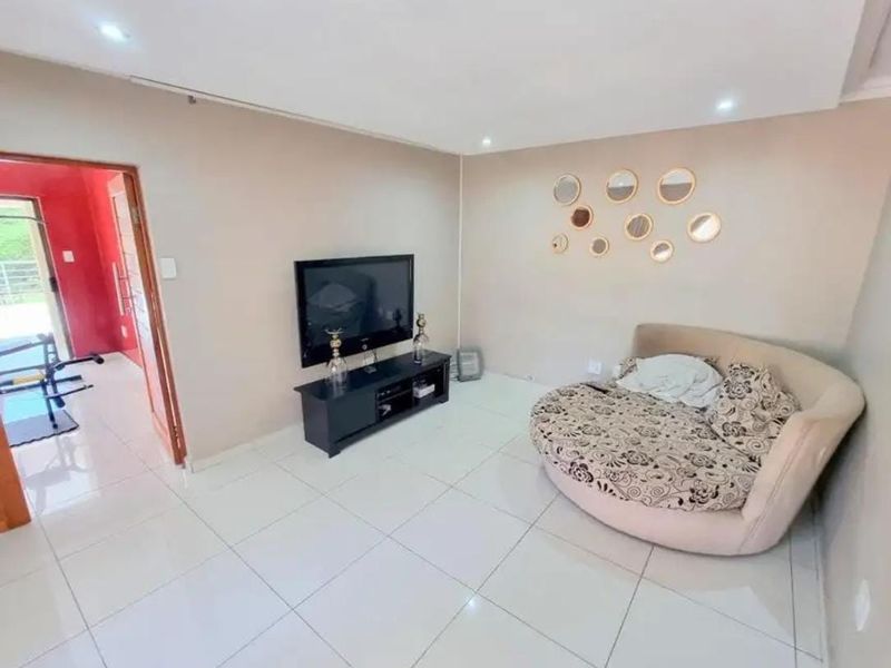 A living room featuring large format floor tiles, recessed ceiling lighting, and a neutral wall finish, with an adjacent doorway leading to an exterior space.