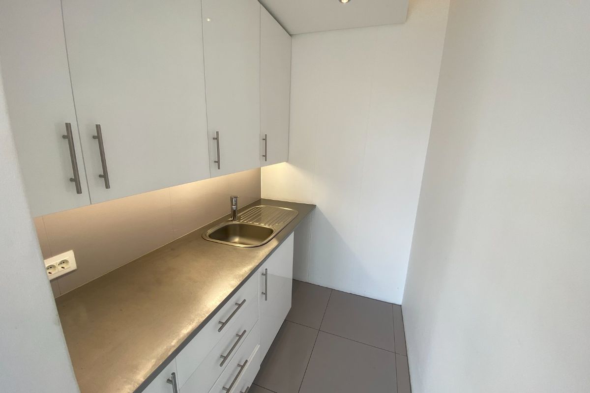 A compact, modern scullery featuring white cabinetry, a stainless steel countertop with an integrated sink, and neutral floor tiling.