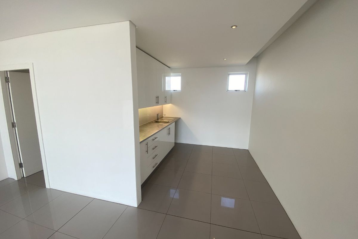 A compact, modern kitchenette featuring white cabinetry, stone countertops, a stainless steel sink, and neutral floor tiling in an open-plan space.