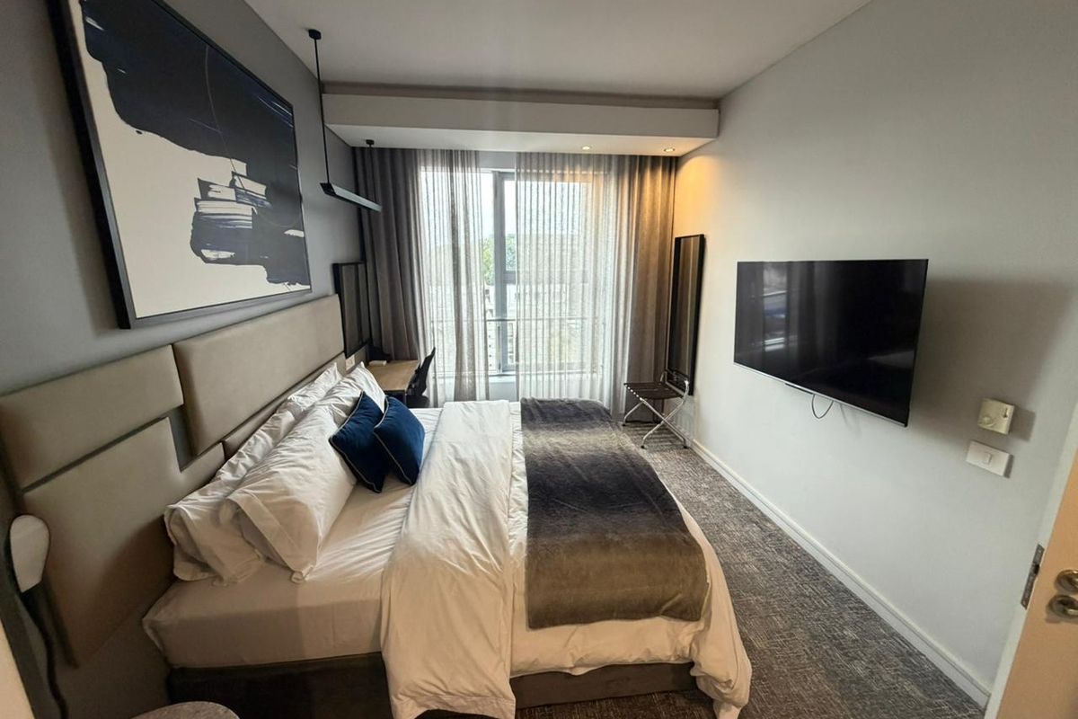 A modern, well-maintained bedroom featuring a built-in headboard, wall-mounted television, and floor-to-ceiling curtains leading to a balcony.