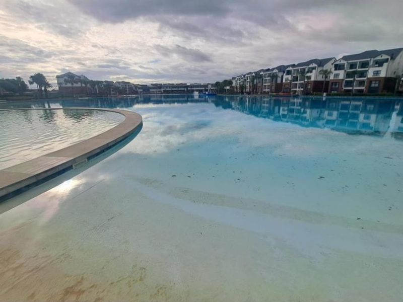 A wide-angle view of a large communal swimming pool area surrounded by modern apartment blocks, featuring a beach-entry design.