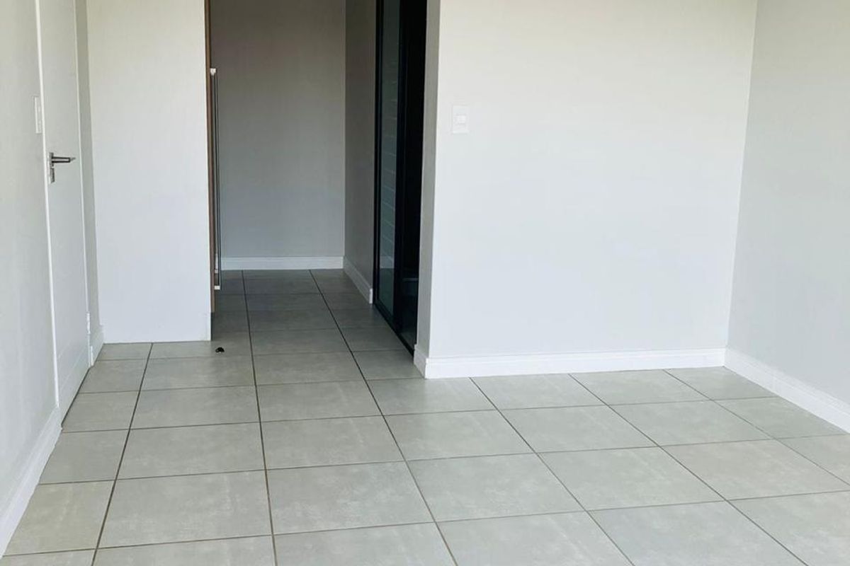 A bright, neutral-toned living area featuring large format floor tiles, coved ceilings, and a clean, minimalist aesthetic leading into a hallway.
