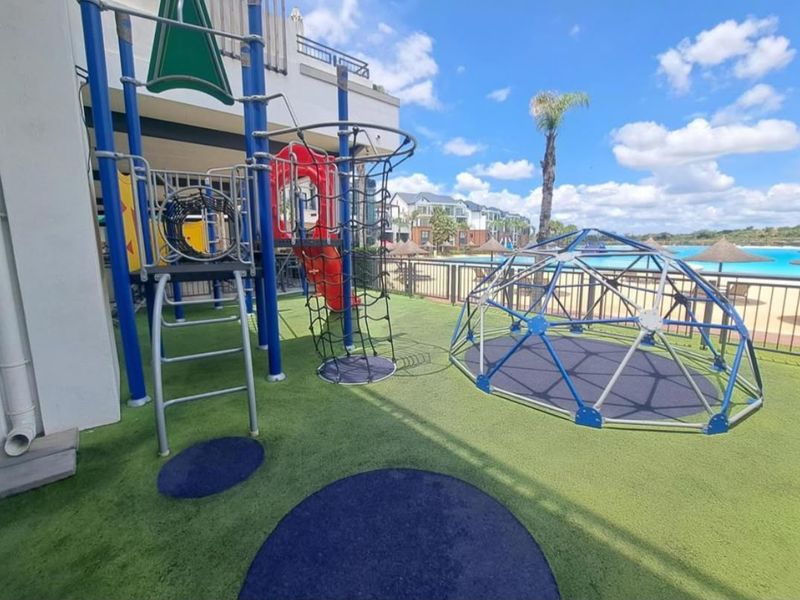 Outdoor communal playground area featuring climbing structures and synthetic turf, situated within a residential apartment complex with pool views.