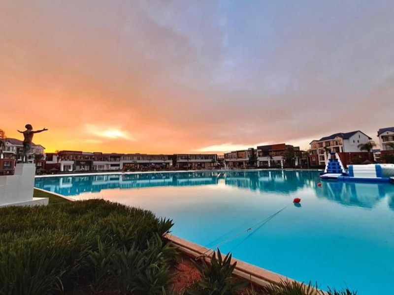 A scenic view of a large communal swimming pool area at sunset, featuring landscaped surroundings and modern apartment building architecture in the background.
