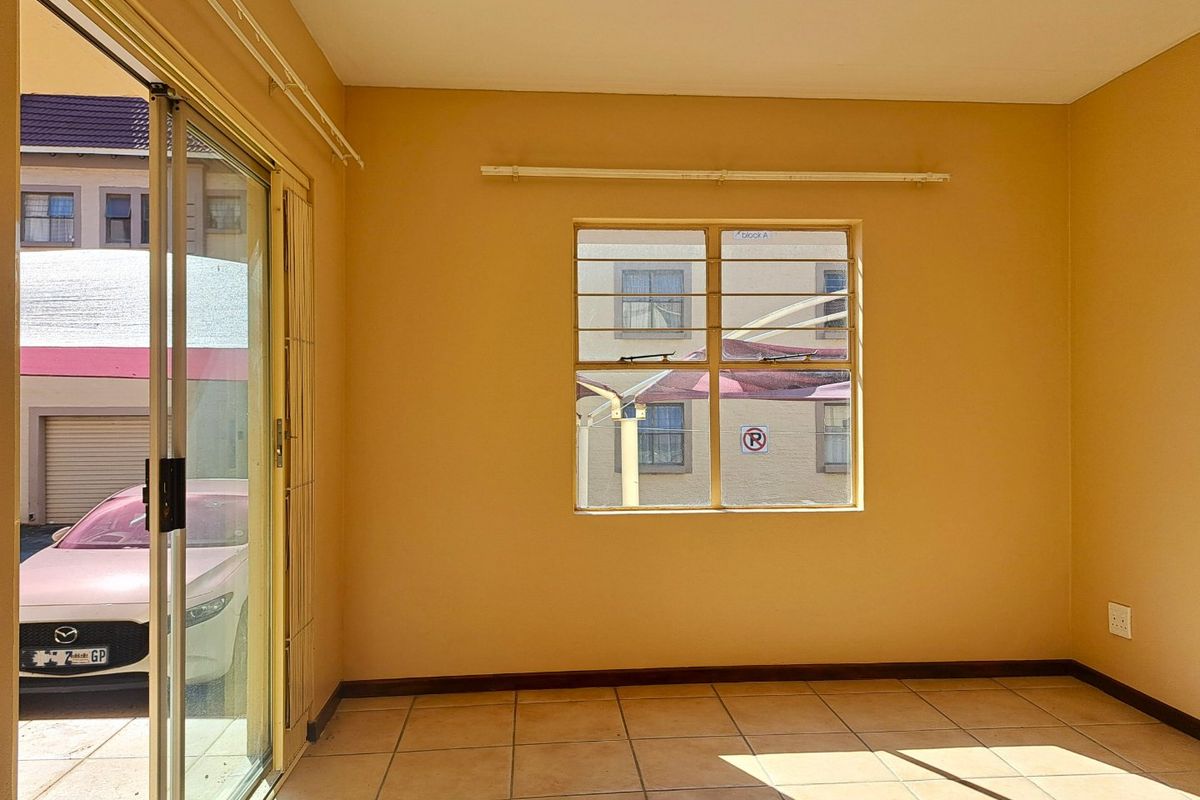 A bright, empty living room featuring tiled flooring, a large window with security bars, and sliding glass doors leading to an exterior area.