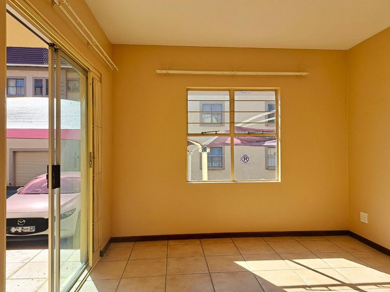 A bright, empty living room featuring tiled flooring, a large window with security bars, and sliding glass doors leading to an exterior area.