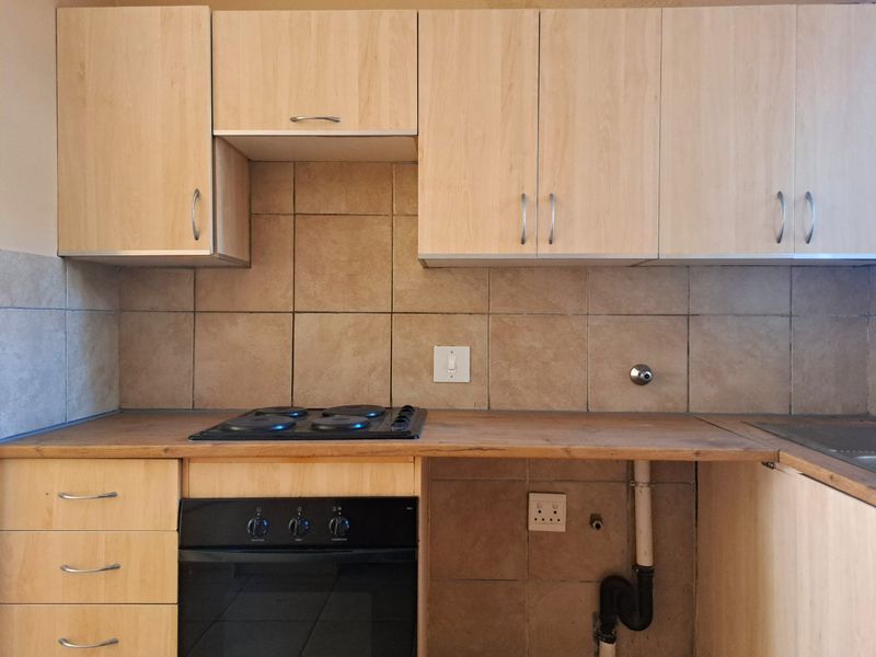 A functional kitchen area featuring light wood-finish cabinetry, a built-in oven, electric hob, and tiled backsplash with dedicated plumbing connections for appliances.