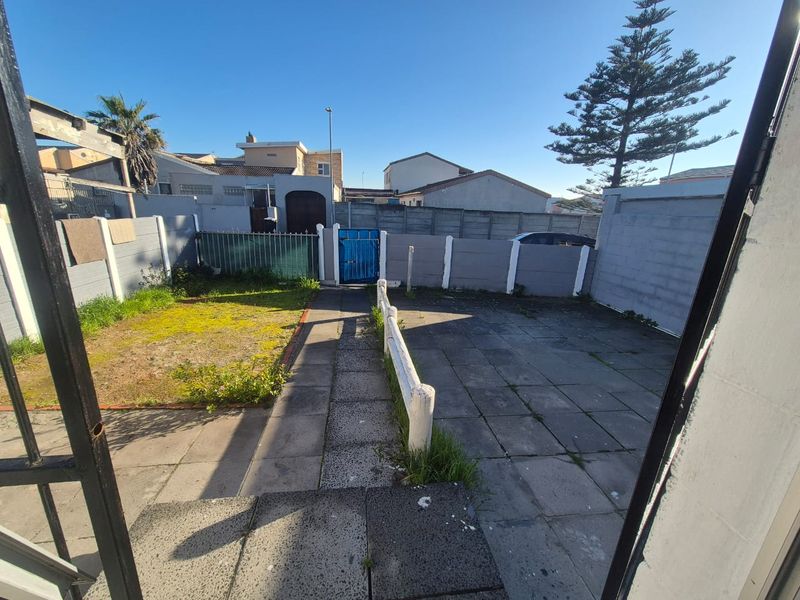 A rear outdoor area featuring a paved patio, a small lawn section, and enclosed boundary walls with a gated access point.