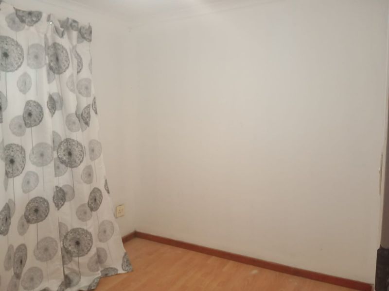A simple bedroom interior featuring laminate flooring and a window with patterned curtains.