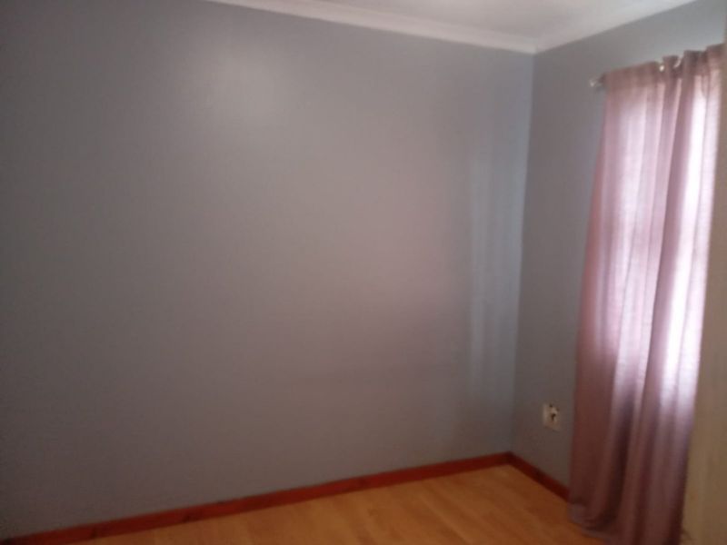 A simple bedroom featuring light-colored walls, wooden flooring, and a window with curtains.