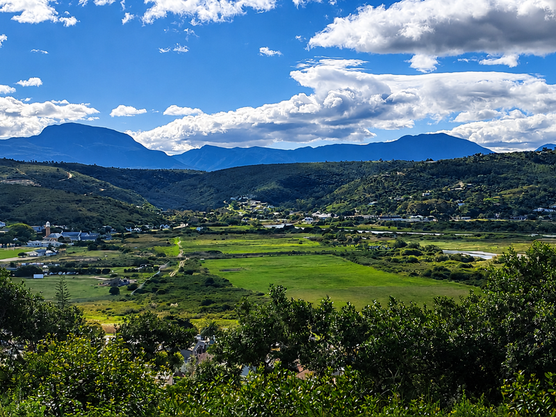A scenic elevated view overlooking a lush green valley, wetlands, and distant mountain ranges under a bright blue sky.