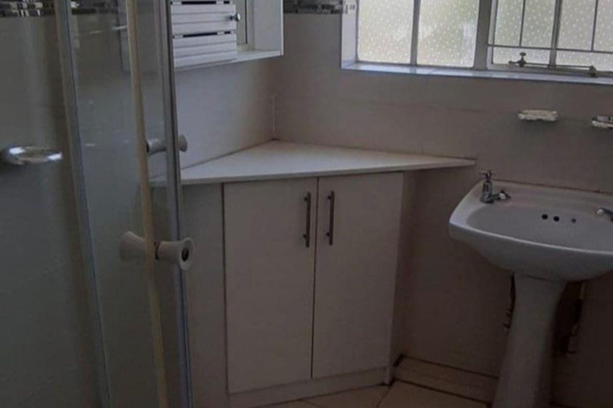 A functional bathroom featuring a corner shower enclosure, pedestal basin, and built-in corner cabinetry with tiled flooring and window security bars.