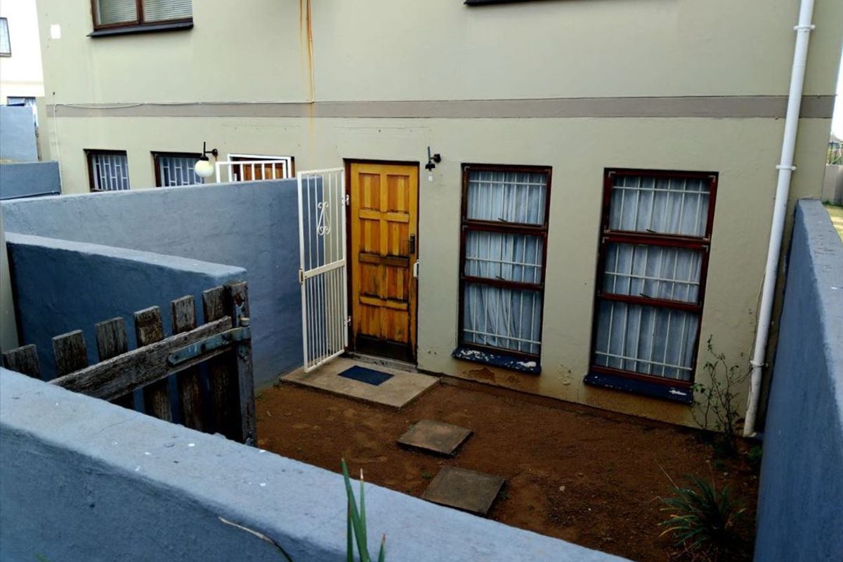 A rear exterior view of a townhouse featuring a small courtyard, security gate, and weathered window frames requiring maintenance.