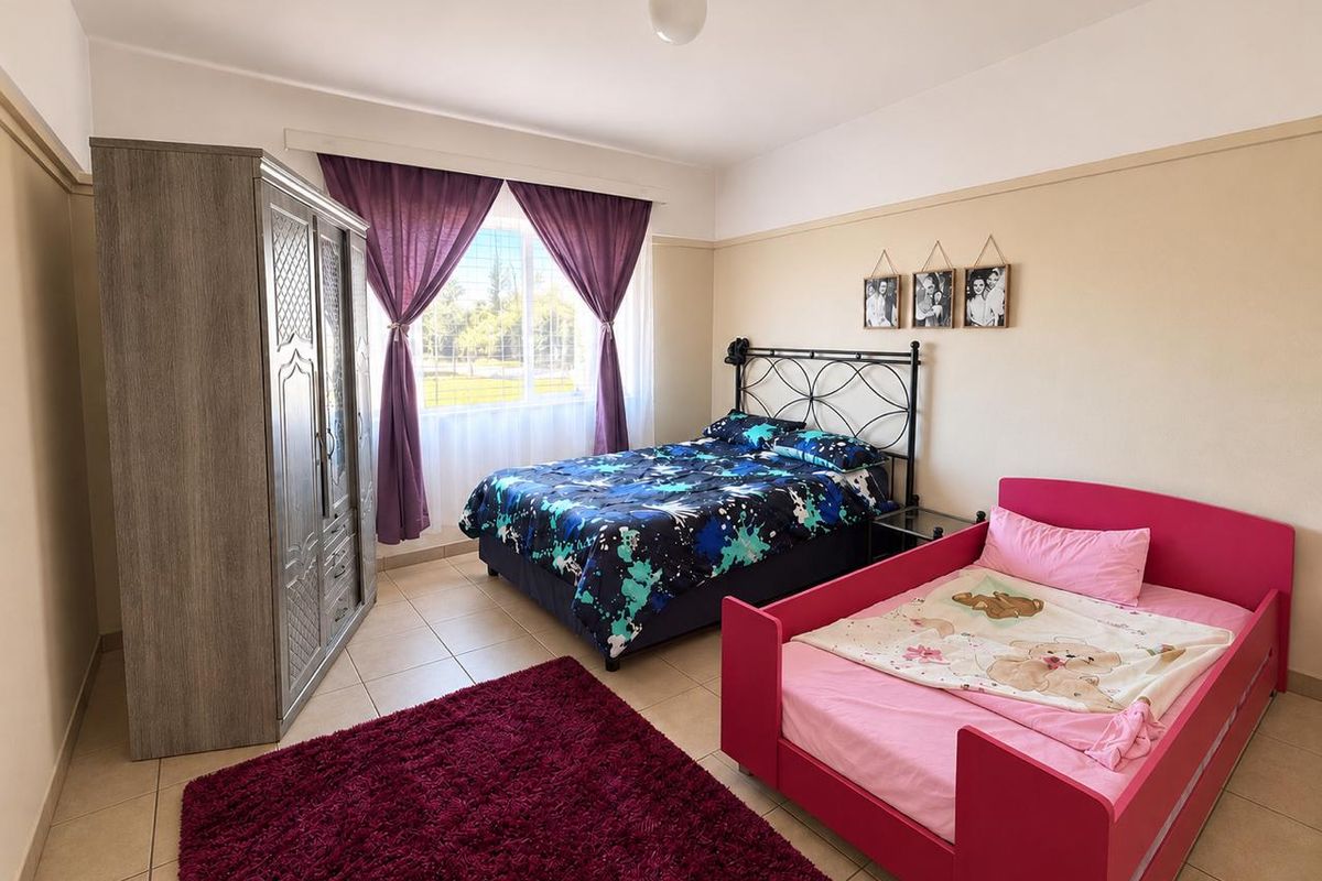 A spacious bedroom featuring neutral wall tones, tiled flooring, and a large window that provides natural light.