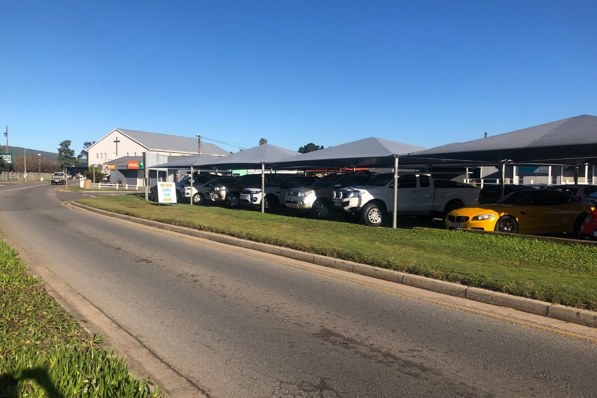 A commercial street-level view of a car dealership featuring multiple shaded parking bays and paved access.