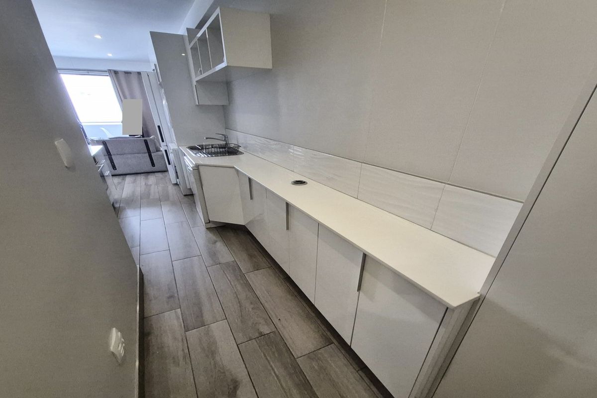 A modern galley-style kitchen featuring white cabinetry, sleek countertops, tiled backsplash, and wood-look floor tiles, leading into an open-plan living area.