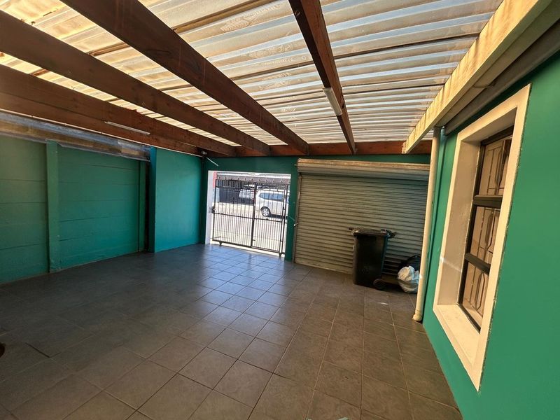 A covered outdoor patio or carport area featuring tiled flooring, a translucent corrugated roof, and secure metal roller shutter door access.