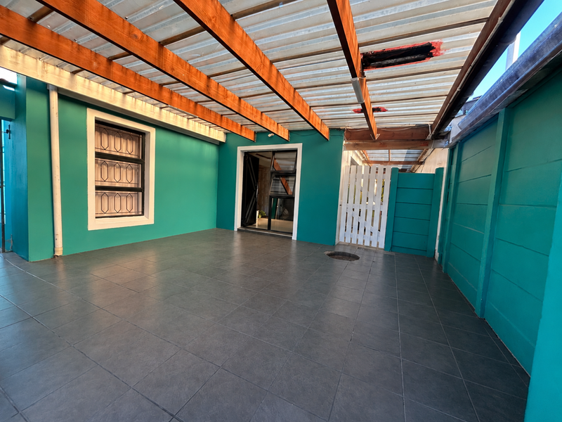 A covered patio area featuring tiled flooring, teal painted walls, and a timber-framed roof structure with a partially damaged corrugated covering.