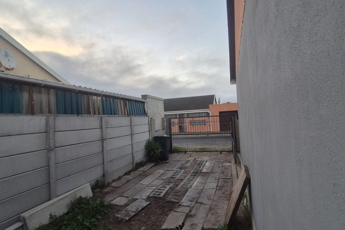 Side exterior area featuring a concrete boundary wall, metal gate, and partially paved ground surface requiring maintenance.