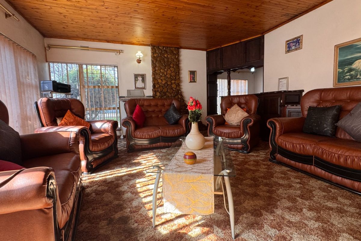 A traditional living room featuring a stone-clad fireplace, wood-paneled ceiling, and large windows with security bars.