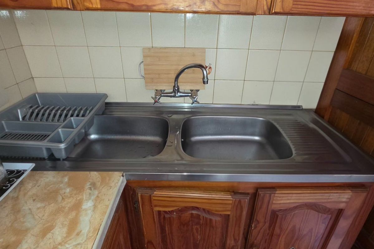 A functional kitchen sink area featuring a double stainless steel basin, wall-mounted faucet, tiled backsplash, and traditional wooden cabinetry.