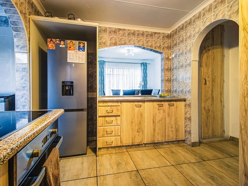 A functional kitchen featuring wood-finish cabinetry, granite countertops, built-in oven and hob, and tiled finishes, with an open archway connecting to the dining area.