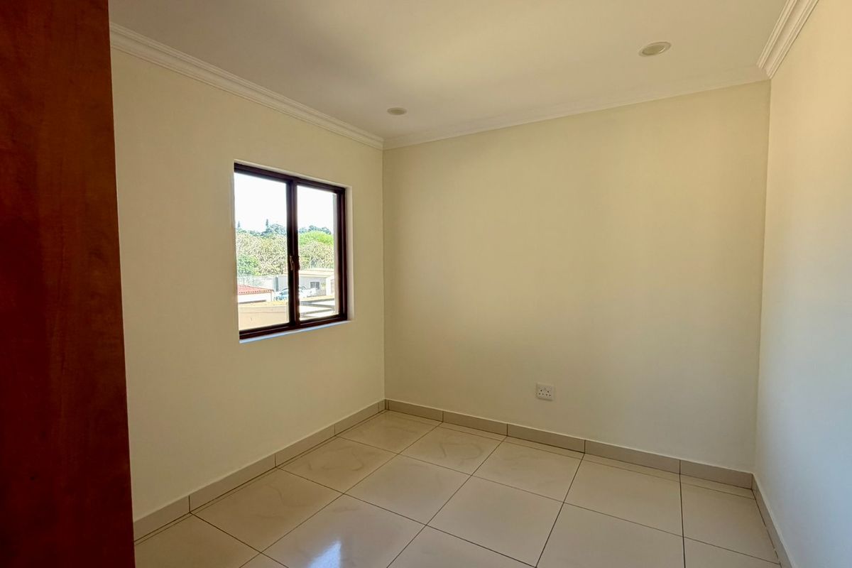 A clean, empty bedroom featuring neutral tiled flooring, recessed downlighting, and a window with an aluminium frame.