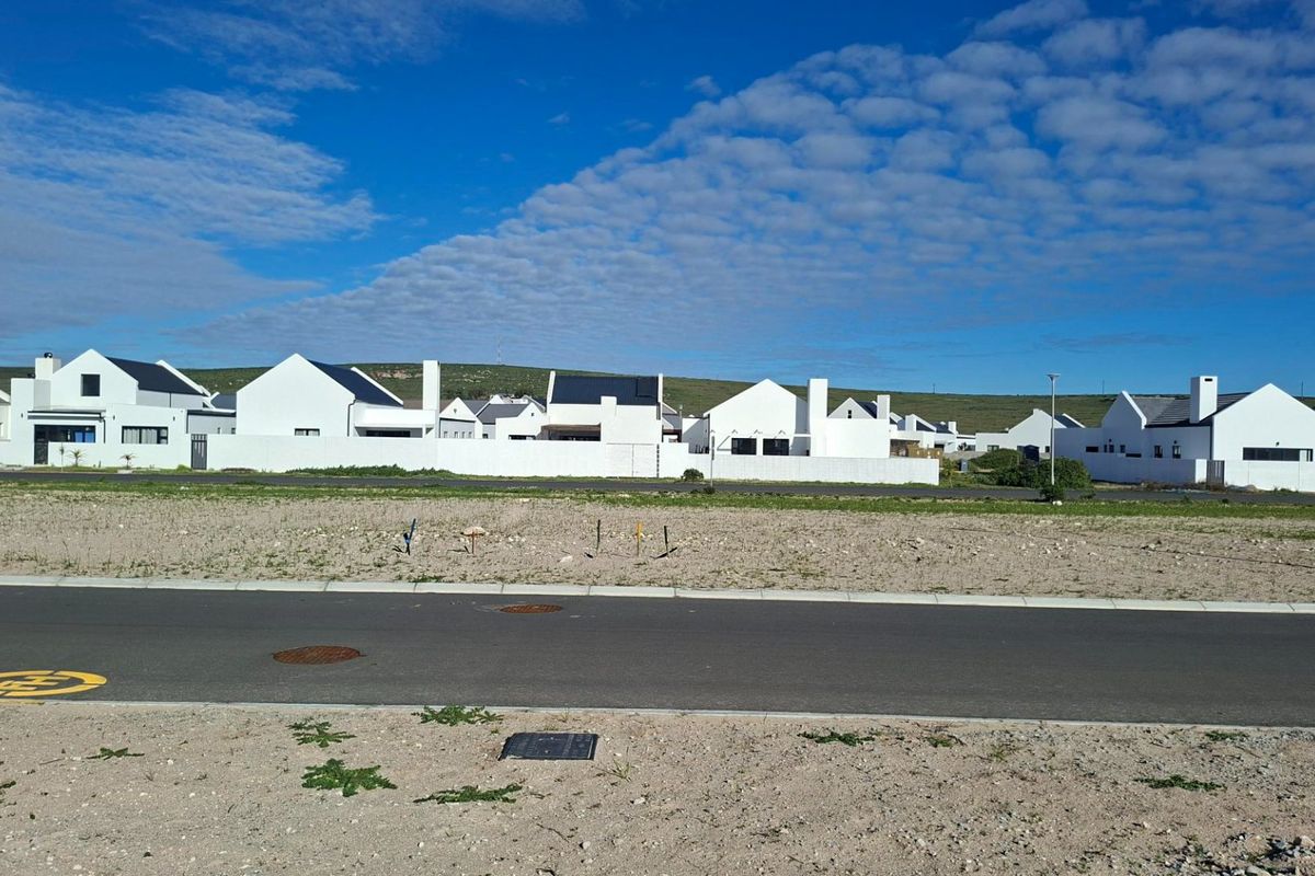 A vacant residential plot situated in a developed estate, featuring paved road access and established surrounding homes.