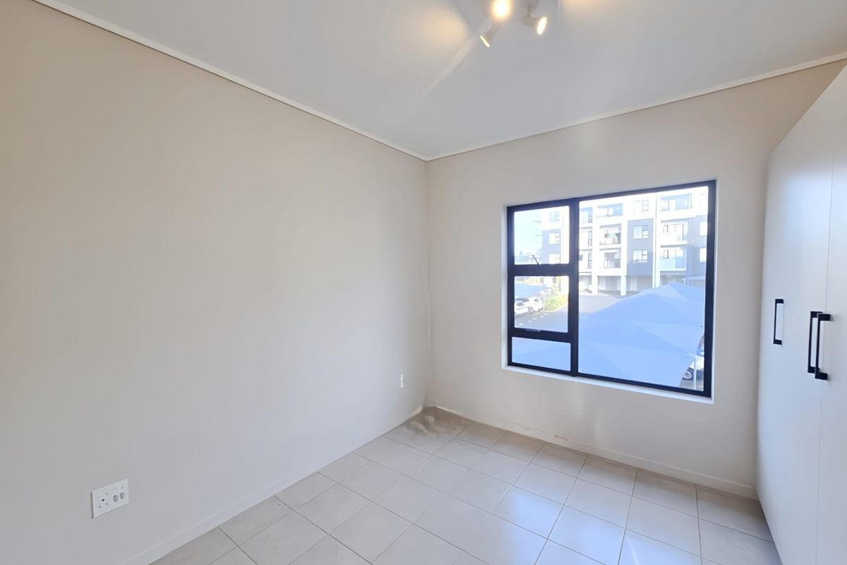 A bright, empty bedroom featuring neutral tiled flooring, a built-in wardrobe, and a large aluminium-framed window providing natural light.