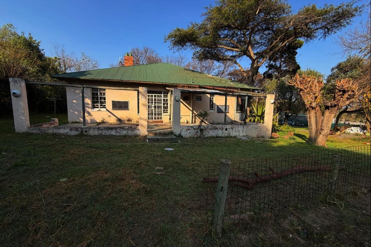 Rear view of a single-story house featuring a covered patio, corrugated metal roof, and a large garden area with mature trees.