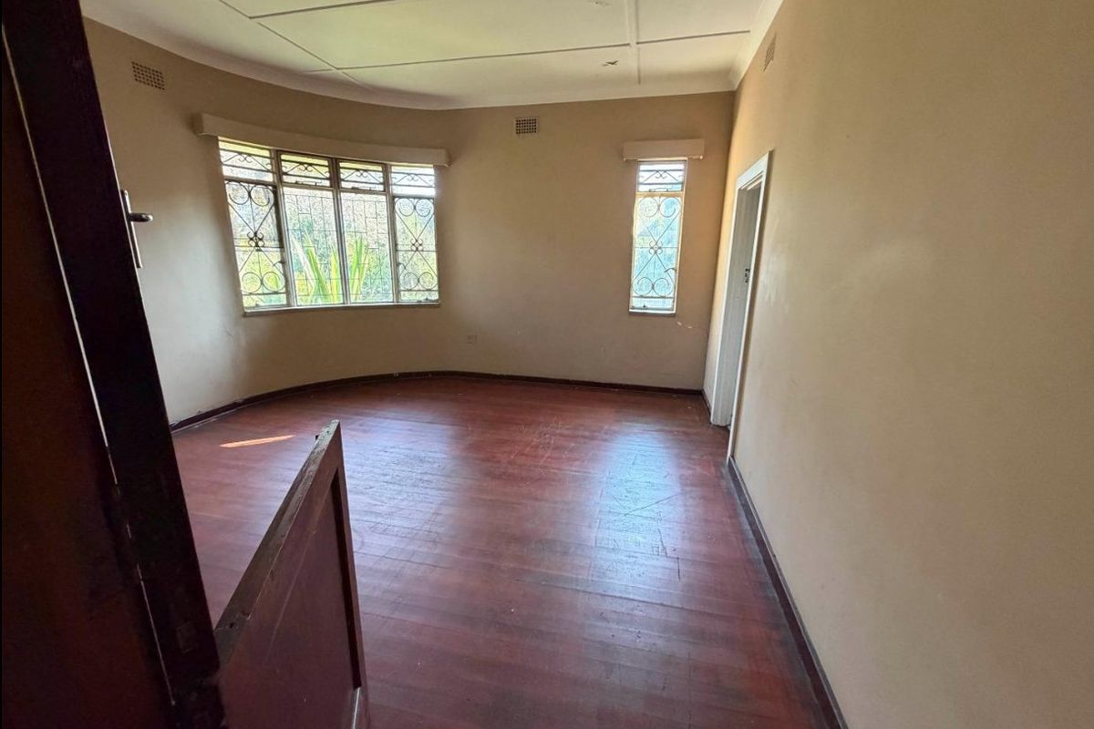 An empty living room featuring original hardwood flooring, a curved bay window with security bars, and neutral wall finishes.