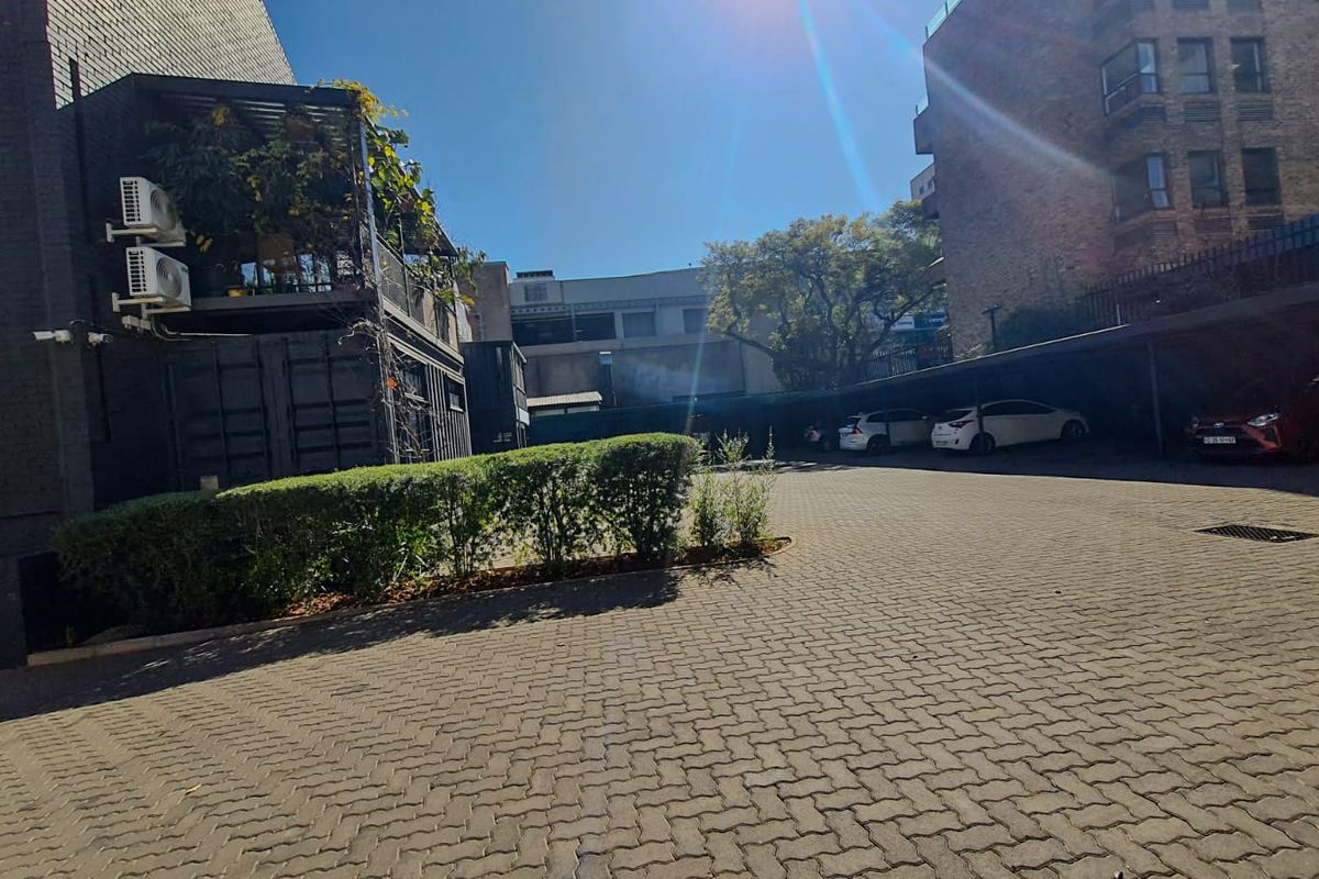 A paved rear parking area featuring a covered carport structure, established hedge landscaping, and external building infrastructure.