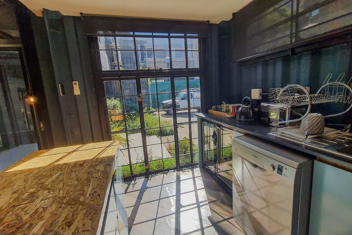 A modern, compact kitchen featuring dark cabinetry, a stainless steel dishwasher, and a large metal-framed glass door providing natural light and garden access.