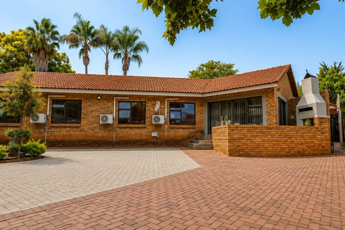 Rear exterior view of a brick house featuring a paved courtyard, built-in braai area, and well-maintained garden surroundings.