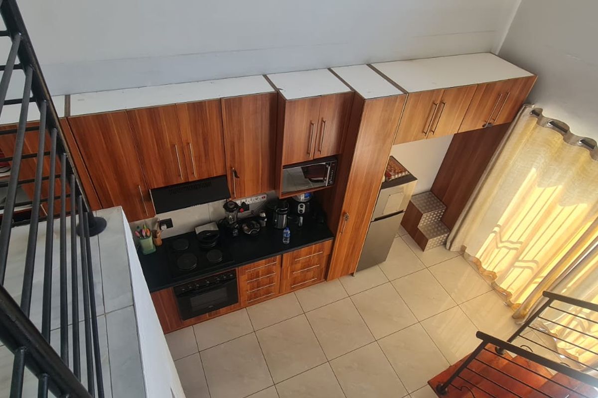 A high-angle view of a compact kitchen featuring wood-finish cabinetry, dark countertops, built-in appliances, and tiled flooring, adjacent to a staircase.