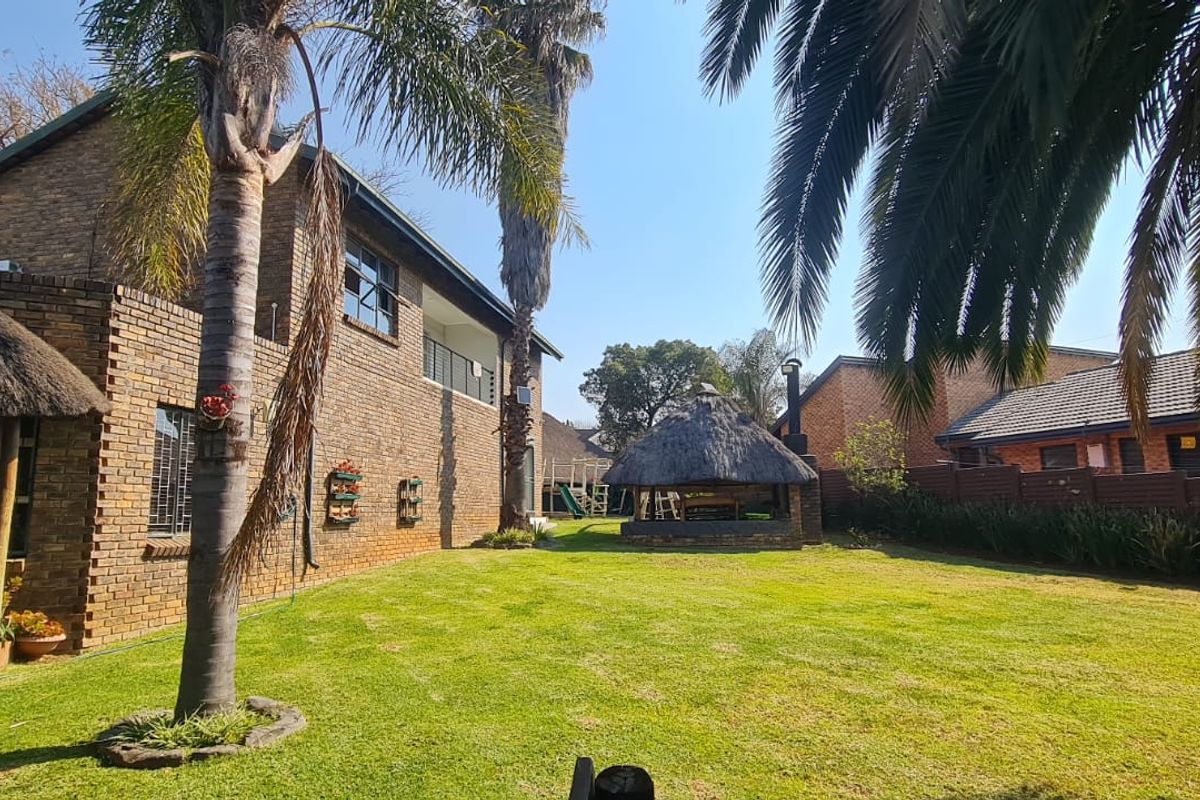 Spacious rear garden featuring a face brick house, a thatch roof lapa with a built-in braai, and established palm trees.