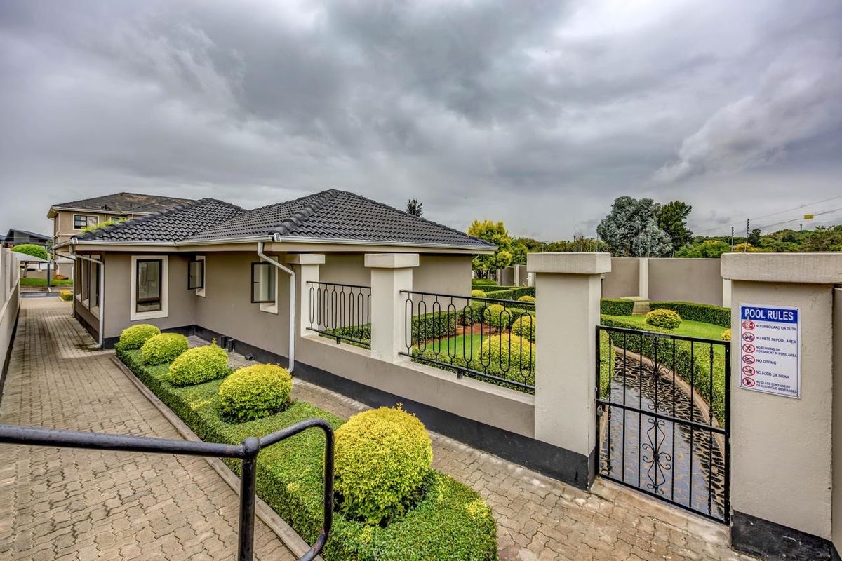 A well-maintained exterior rear area featuring a paved walkway, manicured garden hedges, and secure wrought iron fencing with a gated entrance.