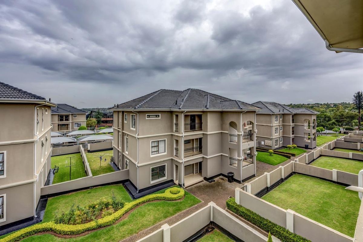 An elevated aerial view of a modern apartment complex featuring multi-story buildings, landscaped garden areas, paved driveways, and covered parking bays.