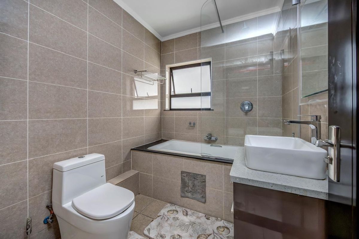 Modern bathroom featuring a built-in bathtub with glass screen, wall-mounted toilet, and a vanity basin set on a stone countertop with neutral wall tiling.