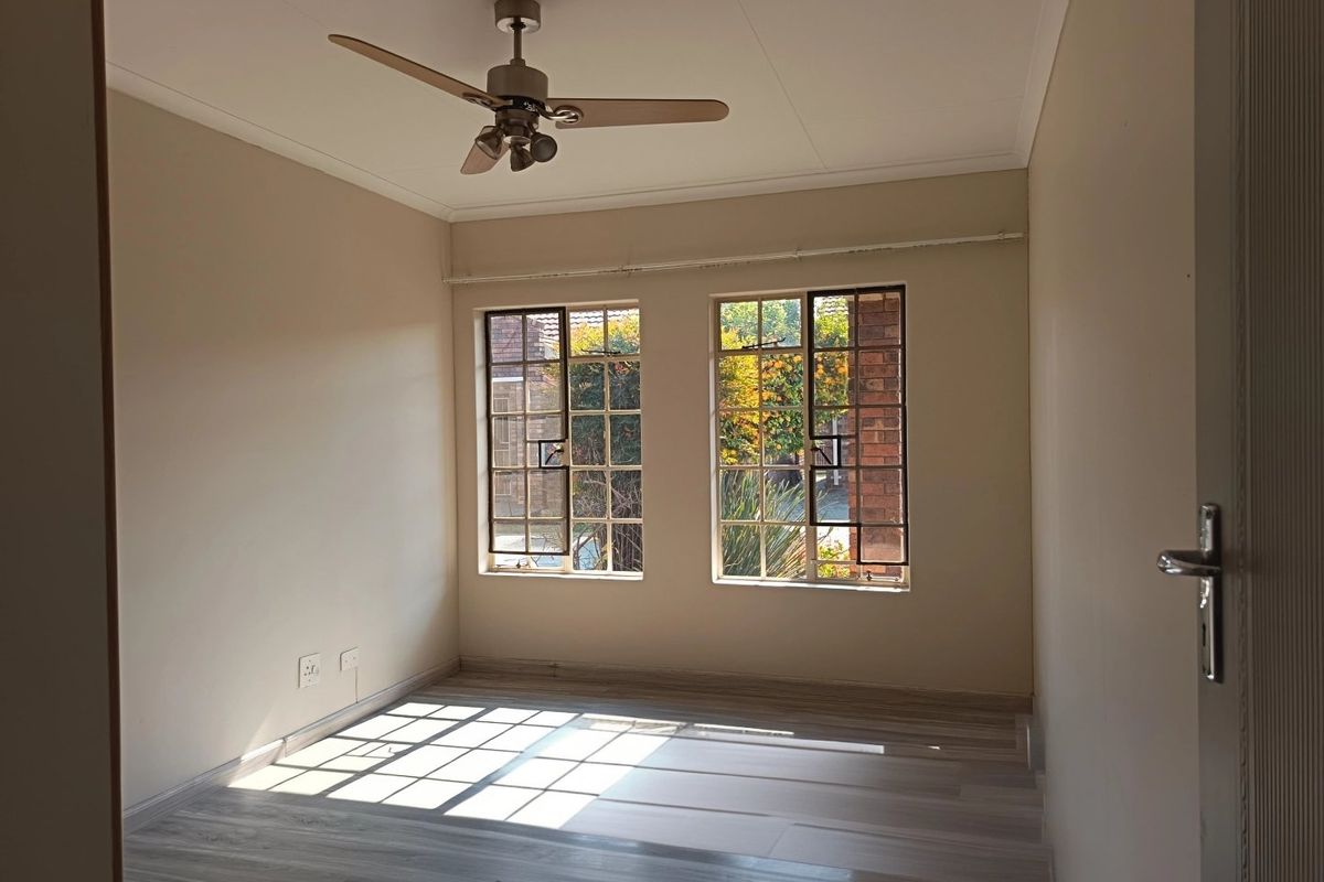 A bright, empty bedroom featuring modern laminate flooring, a ceiling fan, and dual steel-framed windows allowing for ample natural light.