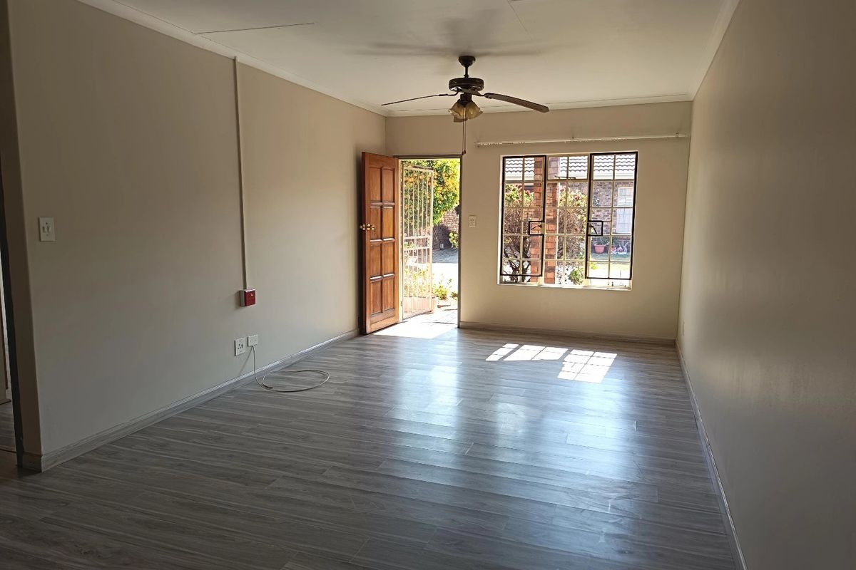 A bright, empty living room featuring modern laminate flooring, a ceiling fan, and natural light through a window with security bars.