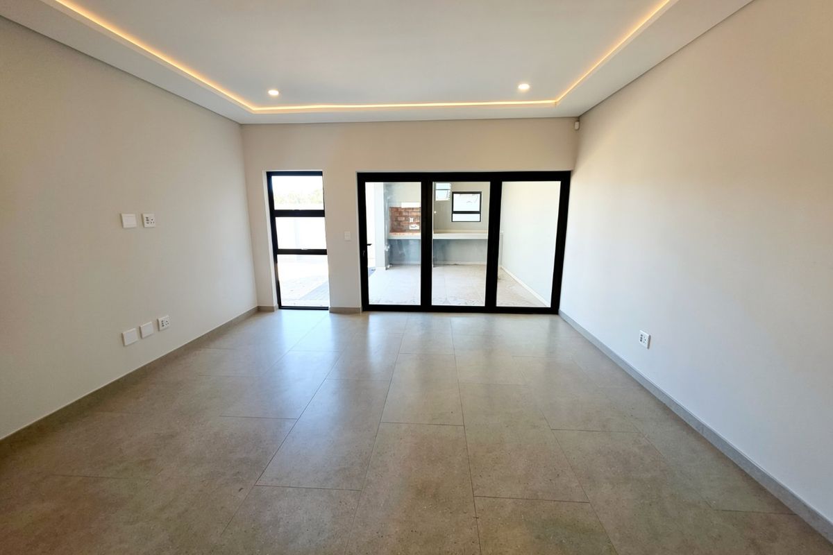 A bright, modern, and empty living area featuring neutral tiled flooring, recessed ceiling lighting, and large aluminium sliding doors leading to an outdoor space.