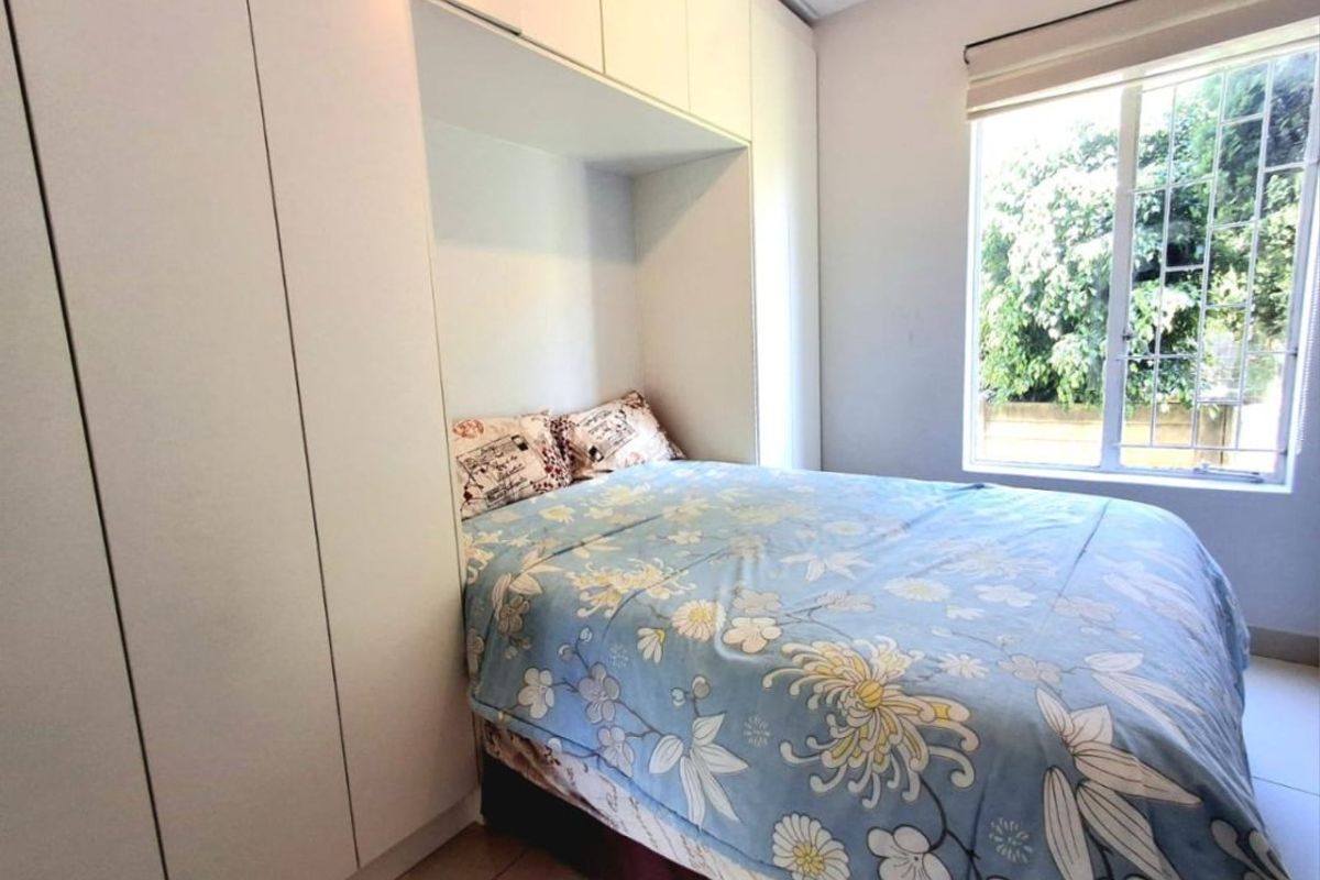A compact bedroom featuring floor-to-ceiling built-in cupboards and tiled flooring, with natural light provided by a window fitted with security bars.