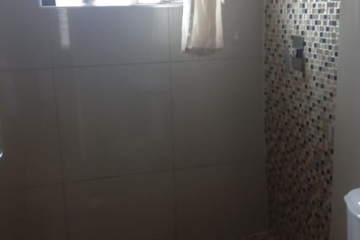 A bathroom featuring a shower area with mosaic tiling, a standard toilet, and a window with security bars.