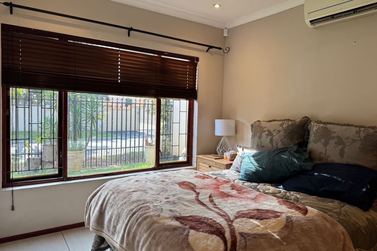 A bedroom featuring tiled flooring, a wall-mounted split-system air conditioner, and large windows fitted with security bars and timber venetian blinds.
