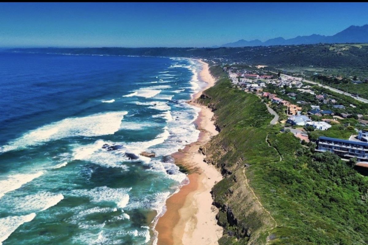 An aerial view of the coastal landscape, showcasing the expansive ocean, sandy beach, and cliffside terrain along the residential area.