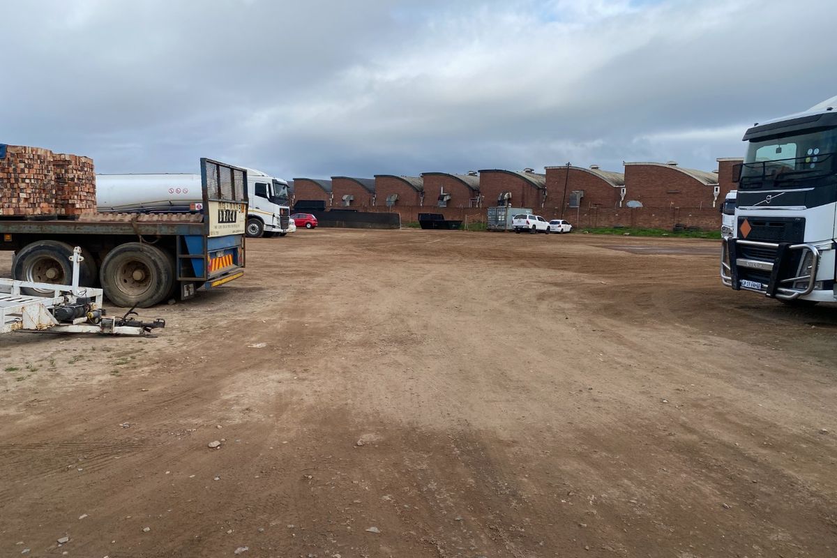 Large unpaved industrial yard area featuring a long warehouse building with brick facade and external ventilation units.