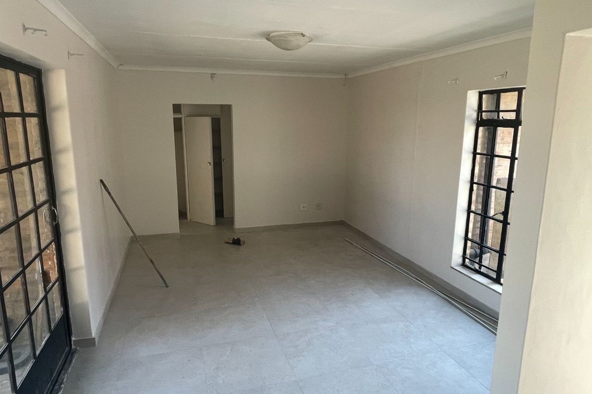 An empty, neutral-toned living room featuring tiled flooring, security bars on the windows, and a secure glass-paneled door leading to the exterior.
