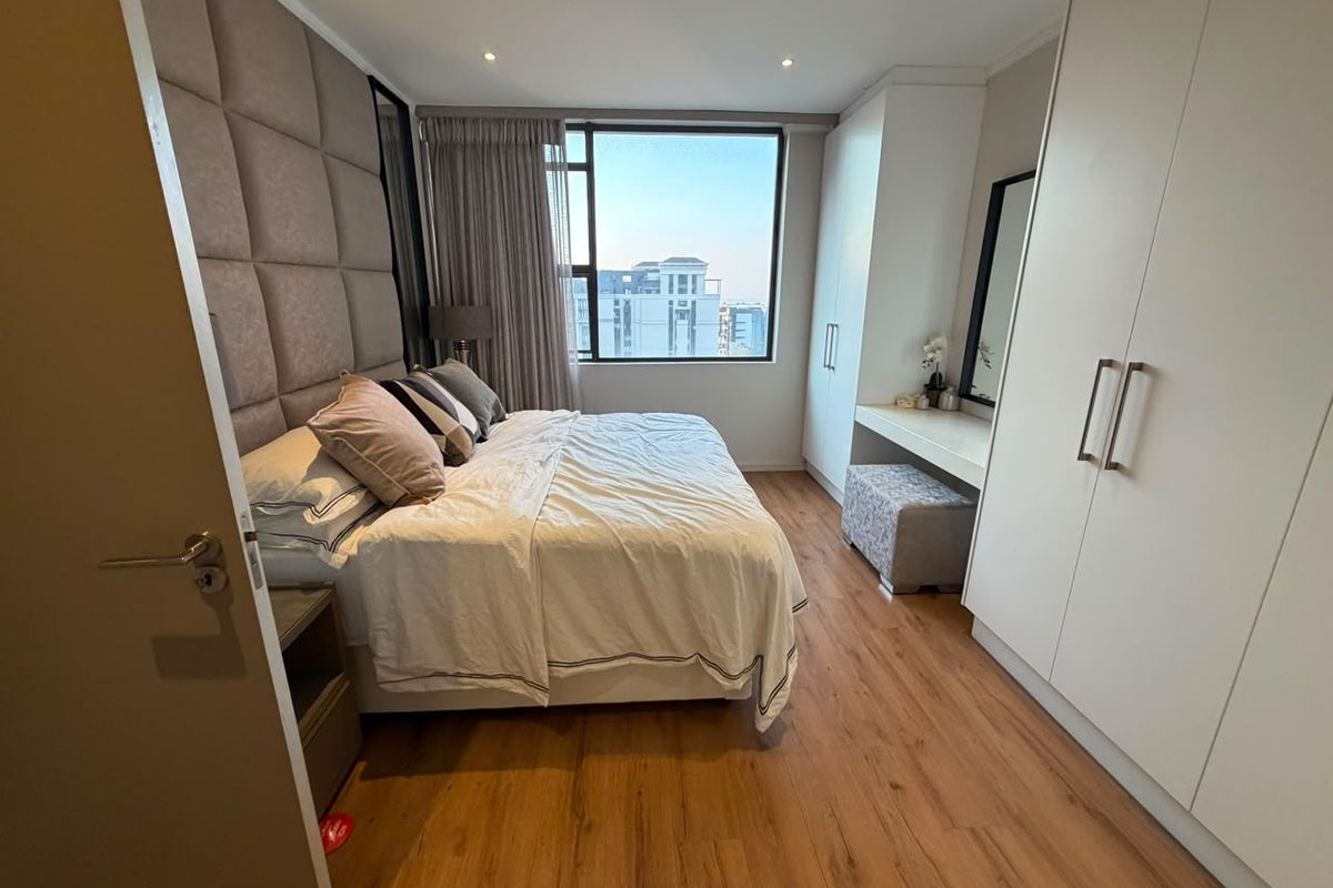 A well-maintained bedroom featuring laminate flooring, built-in cupboards, a custom vanity, and an upholstered headboard with large windows providing natural light.